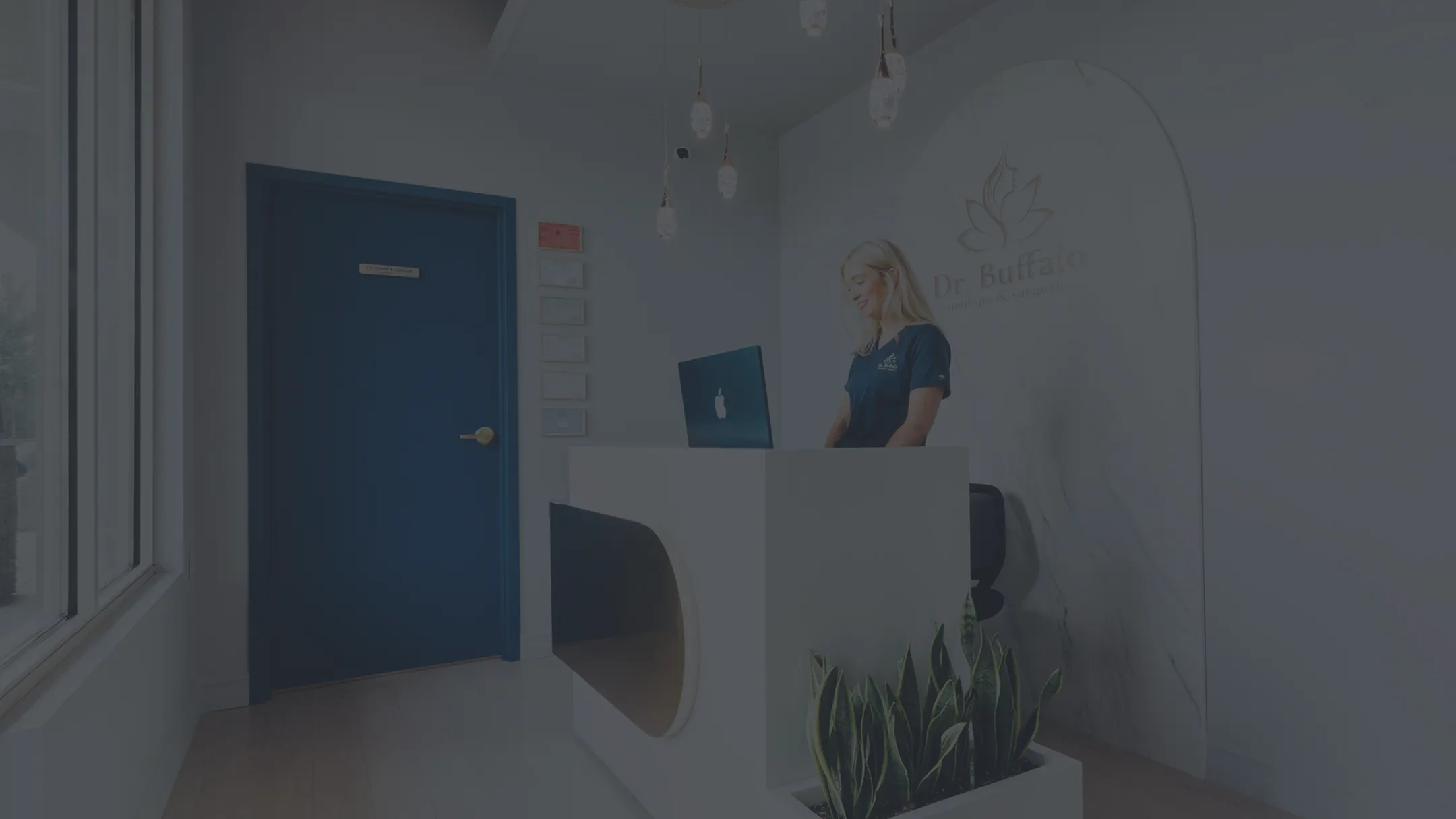 A blonde woman stands behind a modern white reception desk in a brightly lit office. Behind her, a white marble arch features the gold logo and name "Dr. Buffalo," possibly a medical or aesthetic clinic. To the left is a dark blue door.
