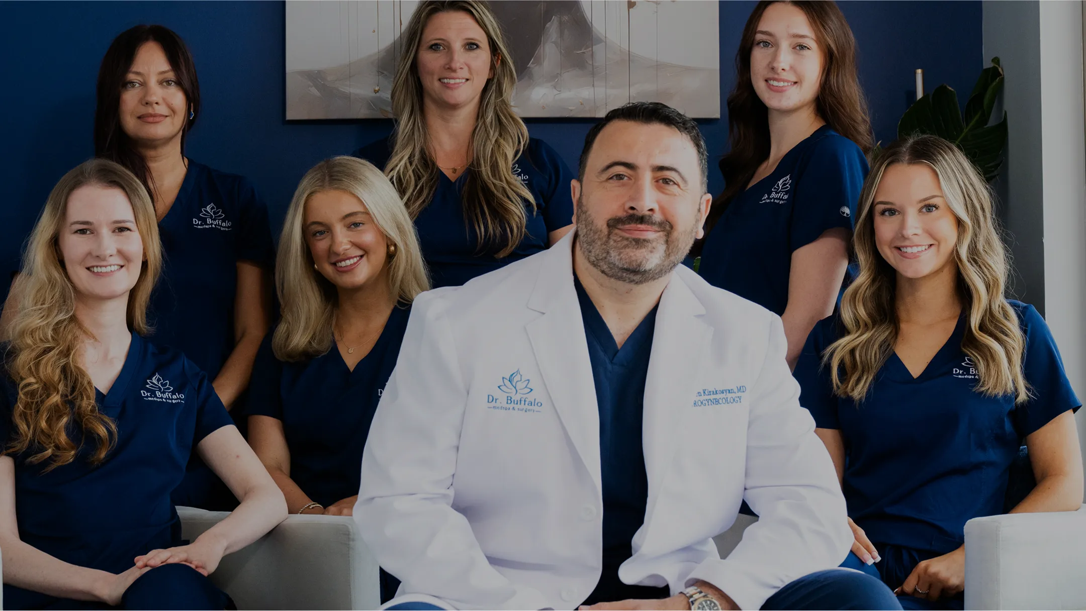 A group photo of Dr. Armen Kirakosyan and his team. Dr. Kirakosyan is seated in the center wearing a white lab coat over blue scrubs. Six women wearing matching dark blue scrubs stand or sit around him, all smiling. The background is a dark blue wall.
