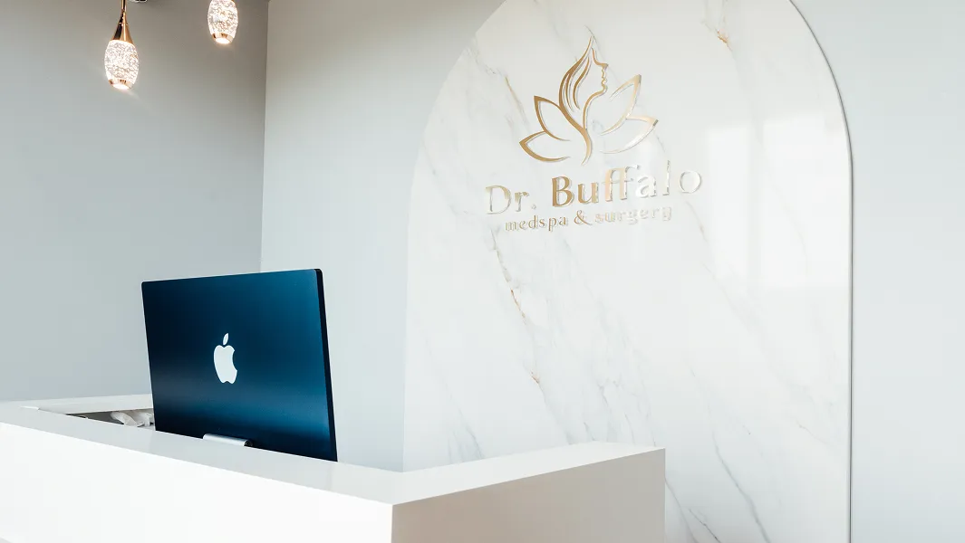 A close-up of a modern reception area with a white desk and a blue computer screen displaying the Apple logo. Behind the desk, a white marble arch features the gold logo and text "Dr. Buffalo medspa & surgery."