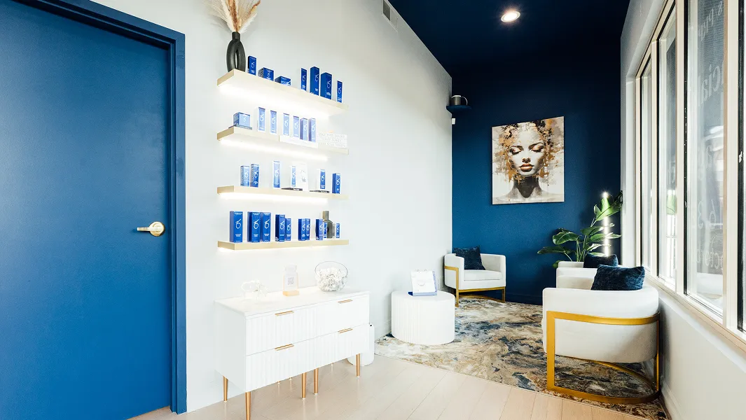A modern, stylish office waiting area. The walls are white and dark blue. A bright blue door is on the left. Floating shelves display blue product bottles. Two white armchairs with gold accents and dark blue throw pillows sit near a window on the right. A painting of a woman's face hangs on the blue wall.