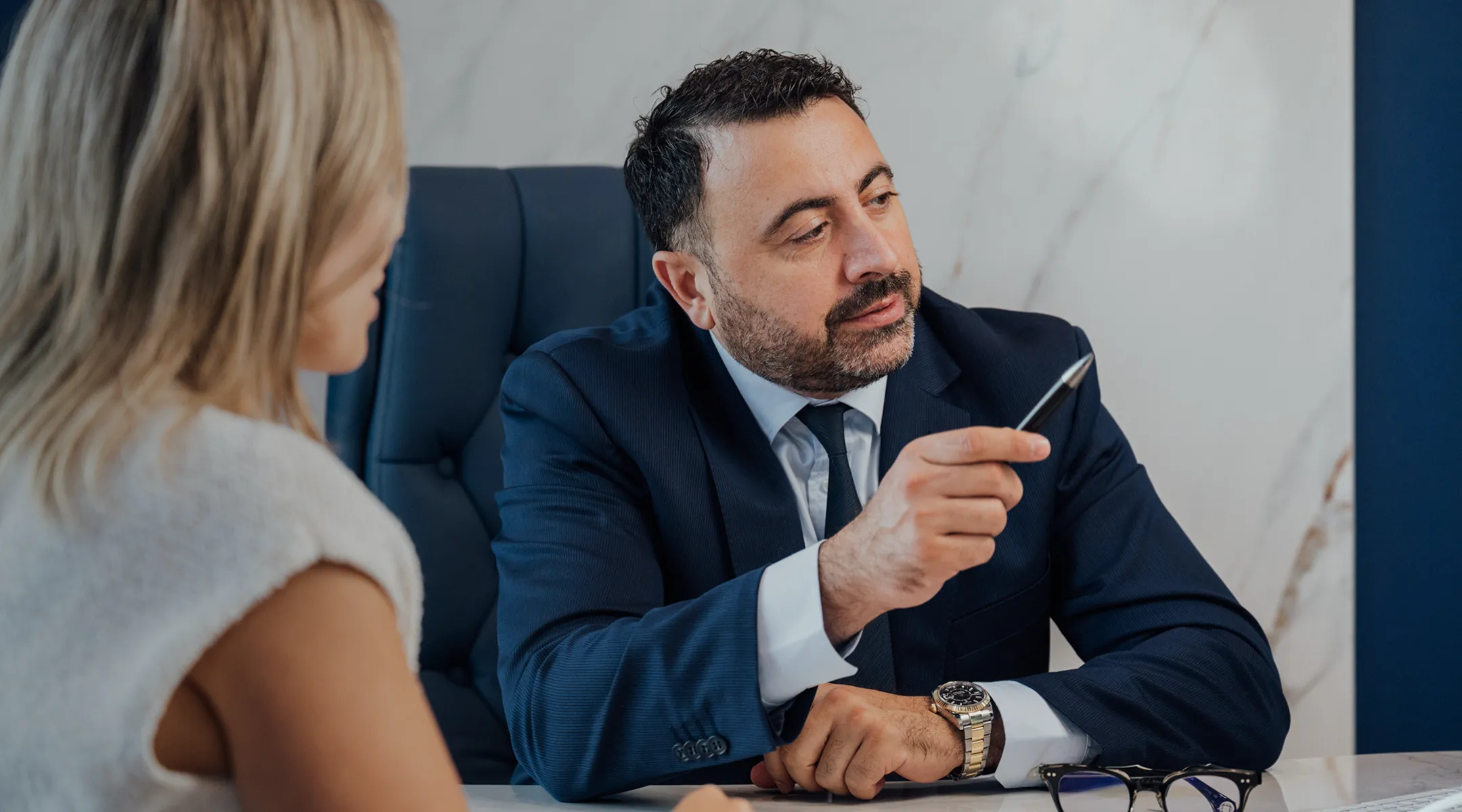 Dr. Kirakosyan, a man in a dark blue suit and tie, sits in a dark leather chair at a white desk, speaking to a blonde woman seated across from him. He is holding a pen and gesturing, with a white marble wall behind him.