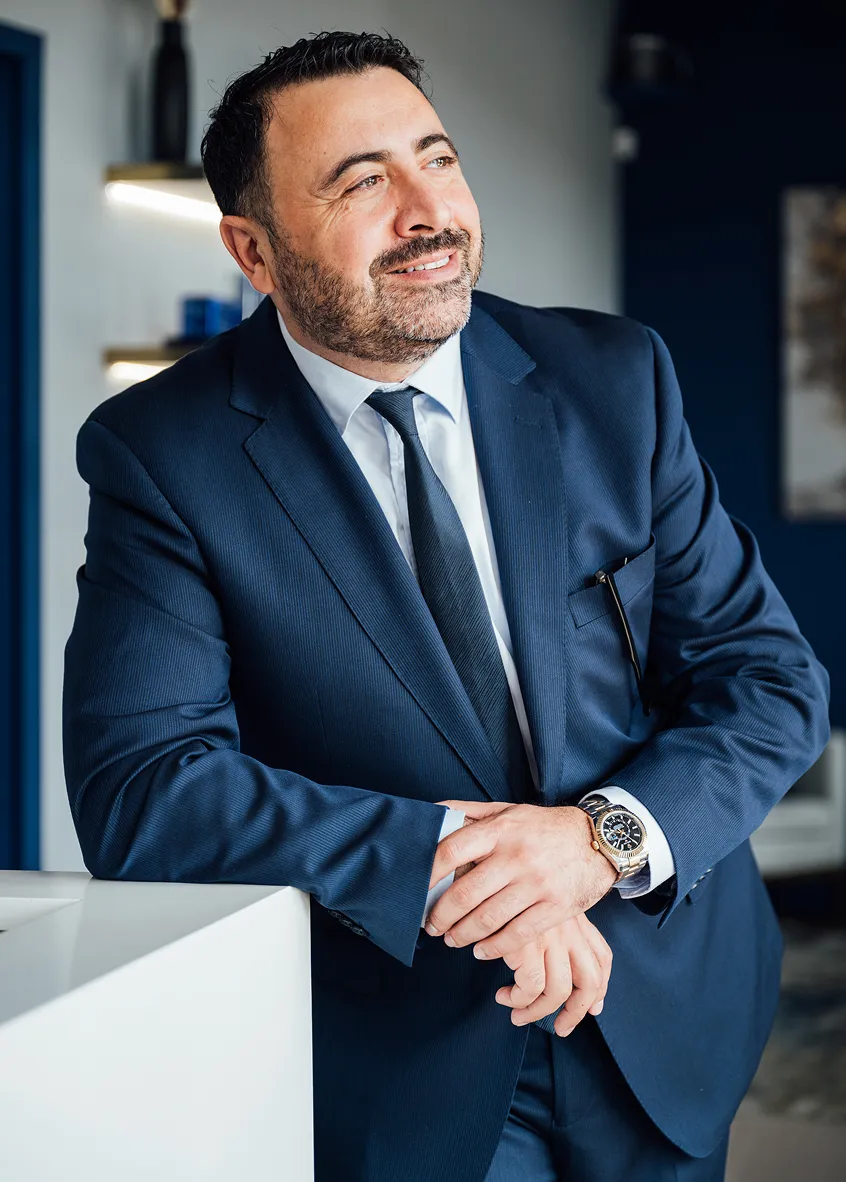 A man in a dark blue suit and tie, Dr. Kirakosyan, is smiling while leaning his arms on a white counter. He is looking up and to the right.