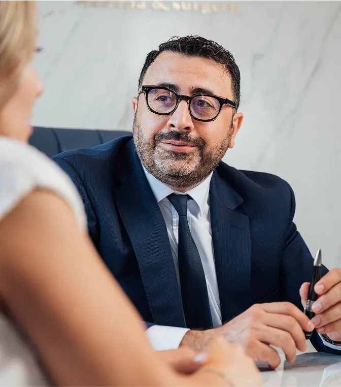 Dr. Armen Kirakosyan, wearing a dark suit and glasses, sits at a desk, looking across at a female patient whose arm is visible in the foreground. He is holding a pen. The background features a gold logo on a white marble wall.