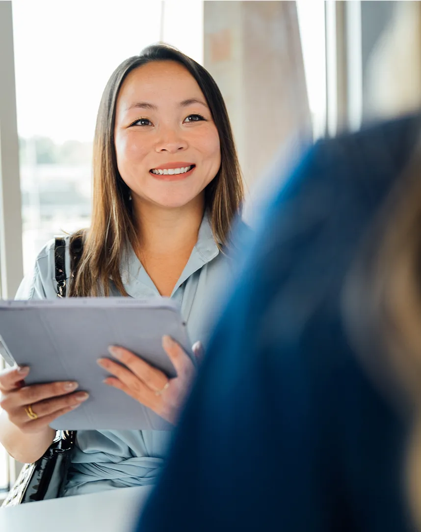 A smiling woman with long dark hair holds a tablet and looks up at a person off-camera, possibly filling out initial paperwork.
