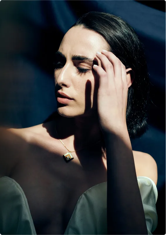 A woman with dark hair and pale skin wears an off-the-shoulder white top and a geometric pendant necklace. She has her eyes closed and her hand touches her temple. Dramatic light and shadow frame her face.