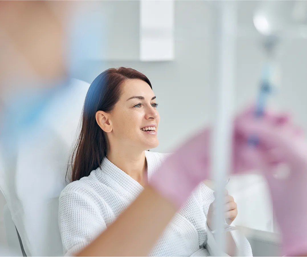 A smiling woman with dark hair is seated and wearing a white bathrobe, looking off-camera. She is receiving IV therapy, and the hands of a technician in pink gloves adjusting the IV line are visible in the foreground.