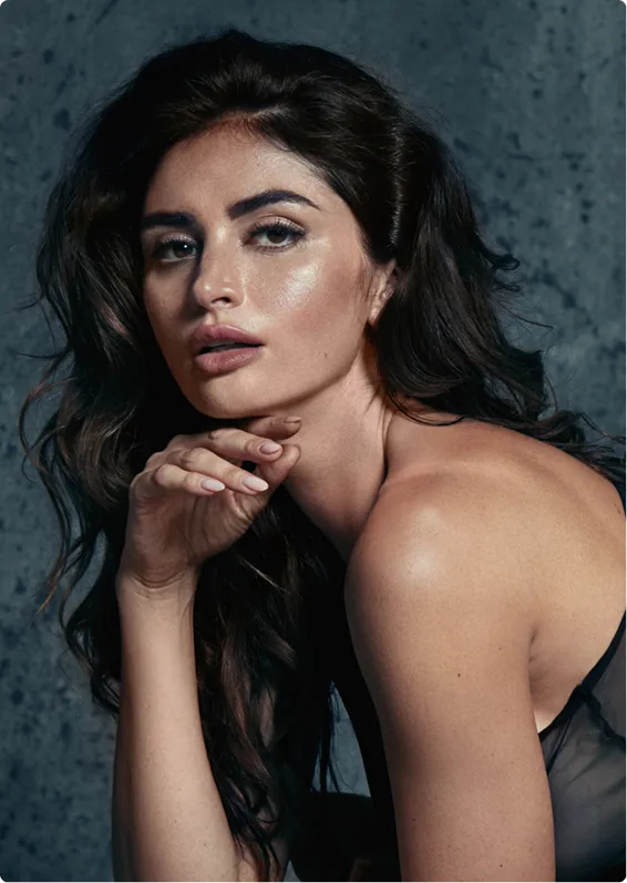 A close up of a beautiful woman with long, dark wavy hair and a full makeup look, wearing a sheer black outfit, leans over against a textured gray wall.