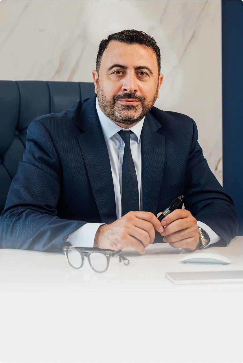 A man in a dark suit and tie, Dr. Kirakosyan, is seated behind a white desk, looking directly at the camera. He is holding a pen, and a pair of glasses rests on the desk in front of him. A white marble wall is visible behind him.