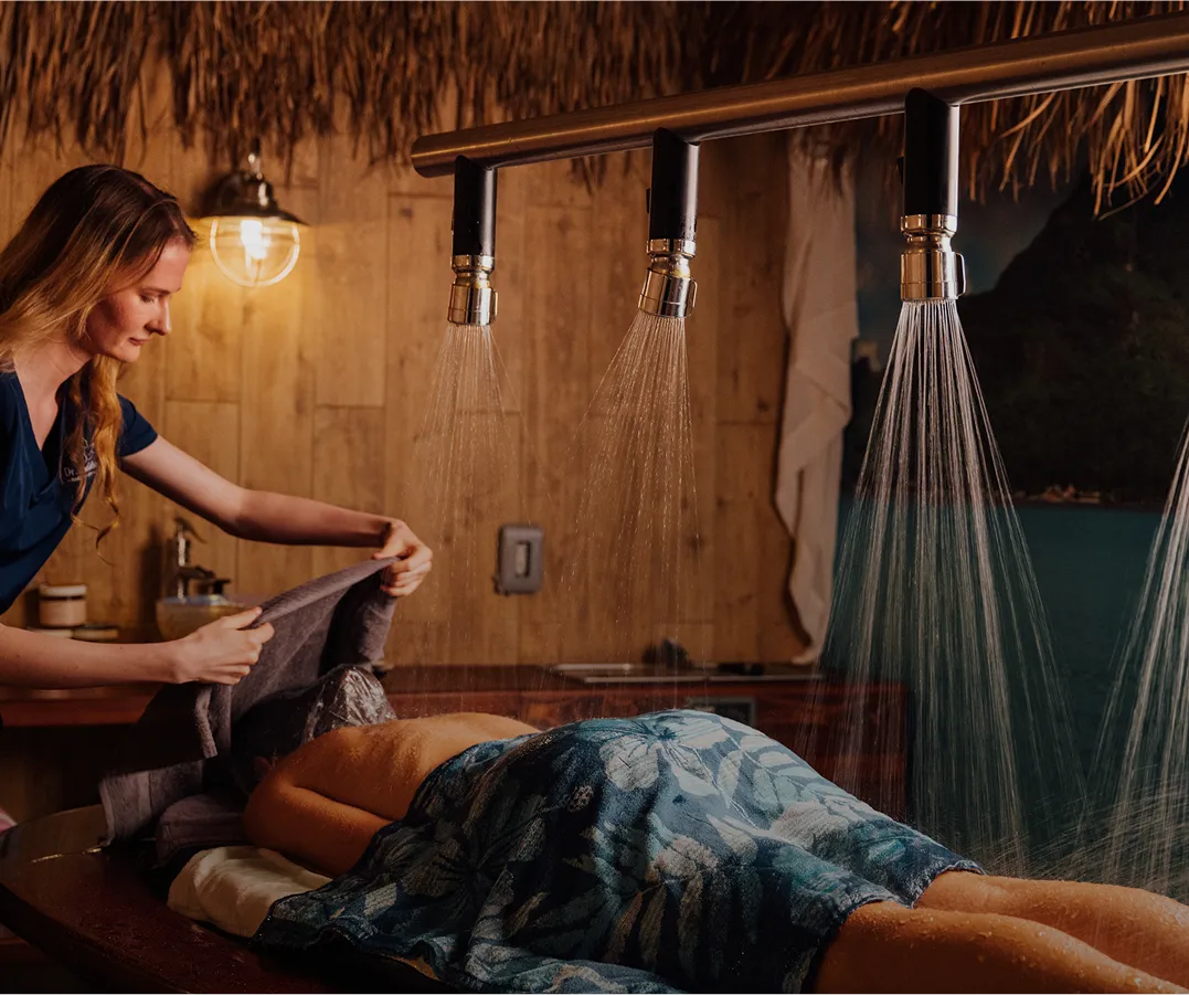 A woman is lying face down on a treatment table, covered with a patterned towel, receiving a Vichy shower treatment. A female technician is standing nearby, preparing another towel. The room has a tropical feel with wood paneling and a thatched roof.
