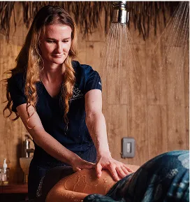 A blonde woman in a dark blue scrub top is giving a client a back massage in a dimly lit, wood-paneled spa room.