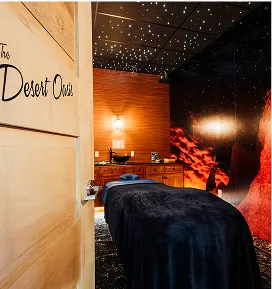 An interior view of a dimly lit spa room named "The Desert Oasis" with a massage bed covered in a dark blue blanket. The ceiling is black with small, white lights resembling stars, and the walls are covered in wood paneling and dark, artistic graphics.