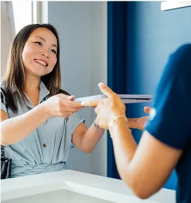 A smiling female client in a light blue shirt is handing documents across a white counter to an unseen person wearing dark blue scrubs.
