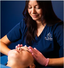 A female technician in dark blue scrubs and pink gloves is performing a facial treatment on a client lying on a table. The technician is gently touching the client's forehead.