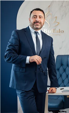 Dr. Kirakosyan in a dark suit and tie, holding glasses, is standing next to a white desk. Behind him, a navy blue wall curves into a white wall featuring a gold logo and the word "Buffalo."