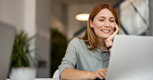 A smiling woman with reddish-brown hair is sitting at a desk, looking at a laptop screen with her chin resting on her hand.