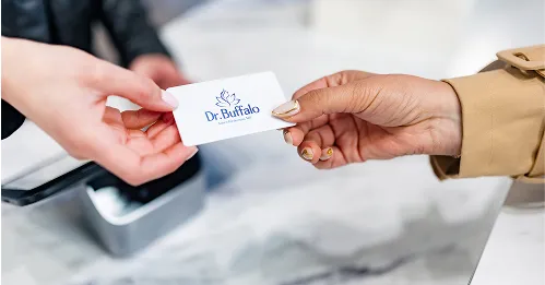 A close-up of two hands exchanging a white card with the blue Dr. Buffalo logo on it. One hand has light skin and pink fingernails, and the other hand has tan skin and gold fingernails. The card is being passed over a countertop next to a payment terminal.