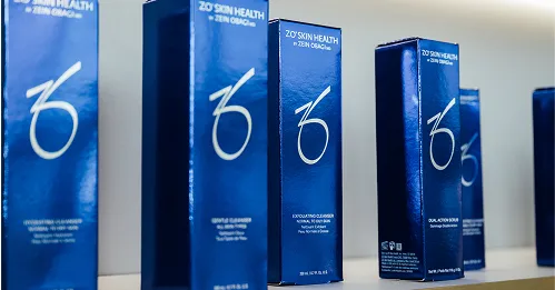 A row of tall, dark blue boxes of ZO Skin Health by Zein Obagi products are displayed on a white shelf. The boxes feature a white stylized 