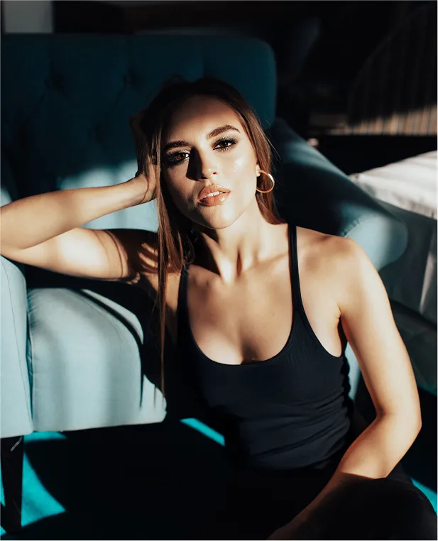 A young woman with long brown hair and gold hoop earrings is sitting down in dramatic, contrasting sunlight and shadow. She is wearing a black tank top and is leaning against a light blue chair.