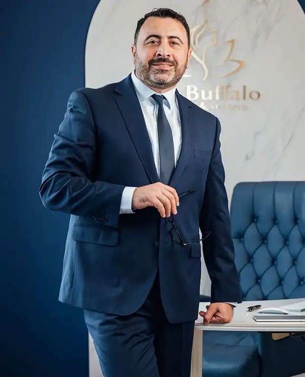 Dr. Kirakosyan in a dark suit and tie, holding glasses, is standing next to a white desk. Behind him, a navy blue wall curves into a white wall featuring a gold logo and the word "Buffalo."