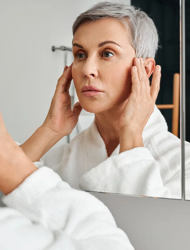 A woman with short, gray hair is looking at her reflection in a mirror. She is wearing a white bathrobe. She is touching her face, and her expression appears somewhat concerned or pensive. The lighting in the bathroom is bright and neutral. The background is a light gray/white bathroom wall. The reflection in the mirror shows a similar-aged woman with a similar expression. - Chemical Peels in Clarence, NY