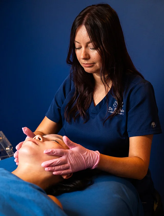 A practitioner wearing blue scrubs and pink gloves gently massages a patient's face during a cosmetic treatment. The background is a solid blue wall. - Co2 Lift in Clarence, NY