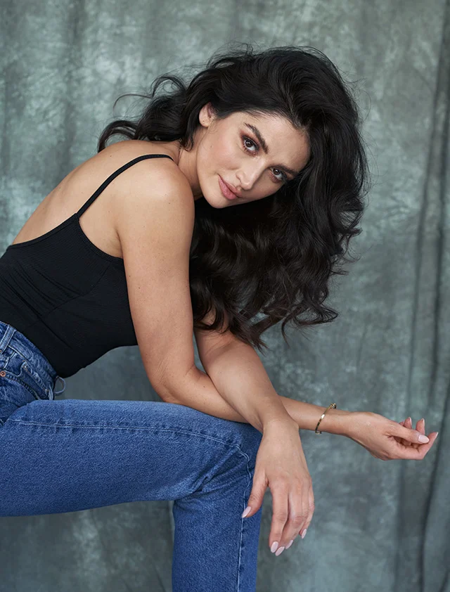 A woman with long, dark curly hair sits against a muted gray-blue textured wall. She wears a black sleeveless top and blue jeans, looking slightly to the left with a neutral, subtly suggestive expression. Soft lighting highlights her features, with the wall serving as a neutral backdrop. - Dermal Fillers in Clarence, NY