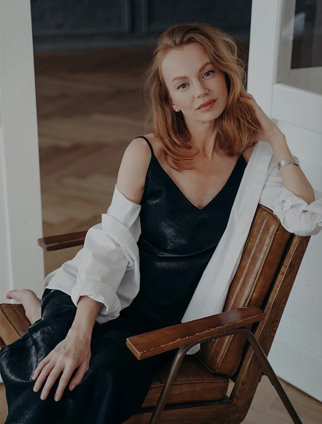 A light-skinned woman with shoulder-length reddish-brown hair is seated, smiling warmly and looking slightly to her left. She wears a dark, likely black, slip dress or top with spaghetti straps. Her joyful expression is complemented by soft, natural lighting. The background shows a light-colored wall and windows, creating a cozy, inviting atmosphere. - Dr. Buffalo Hair Restoration in Clarence, NY