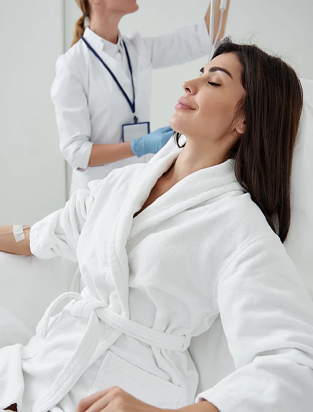 A woman with her eyes closed and a relaxed expression is receiving an IV drip while seated in a chair. She is wearing a white robe and a medical professional is adjusting the IV in the background. - IV Therapy Services in Clarence, NY