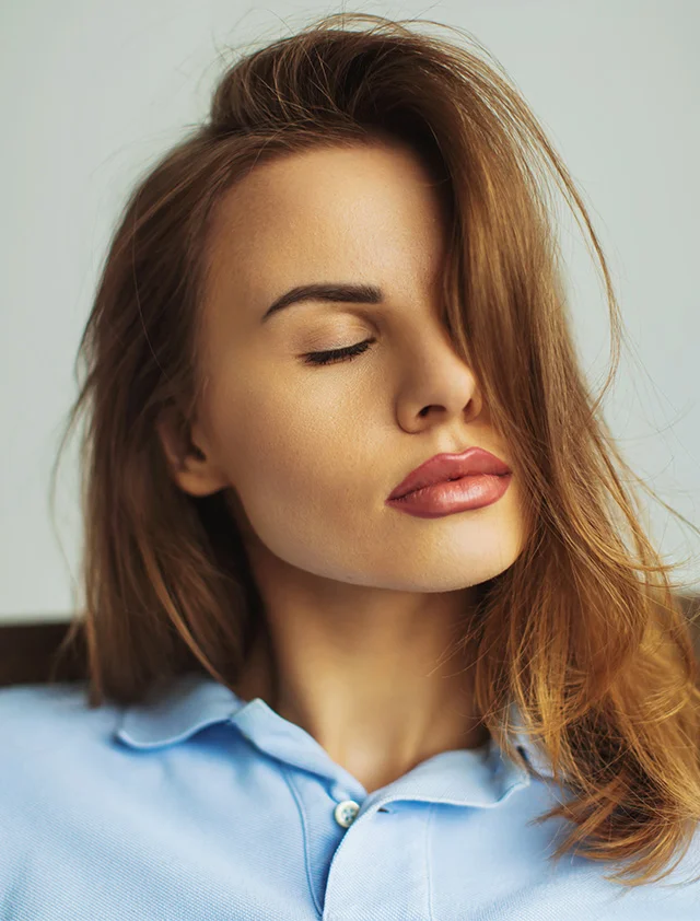 A close-up of a woman with auburn hair covering one side of her face, eyes closed, and wearing a light blue collared shirt. - Lip Fillers in Clarence, NY