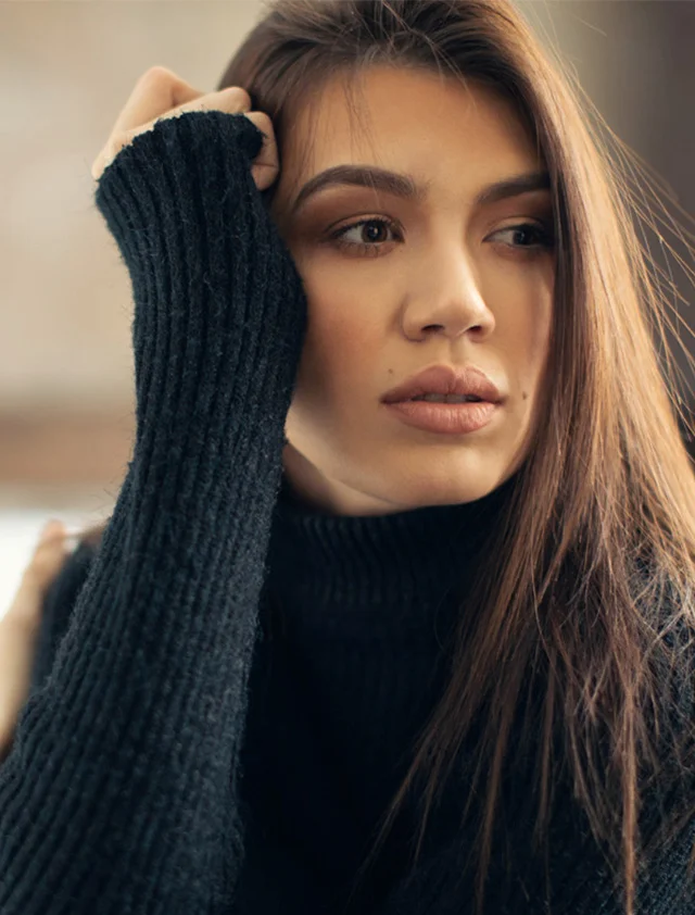 A close-up of a woman with long brown hair wearing a black turtleneck sweater, looking off to the side with her hand resting on the side of her face. - Liquid Facelift in Clarence, NY