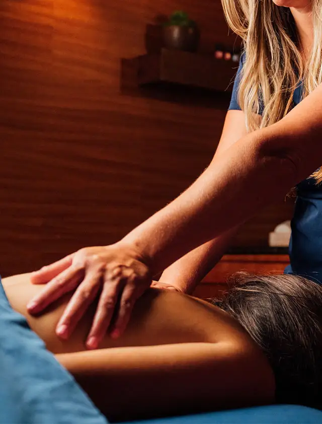 A blonde woman in a dark blue scrub top is giving a client a back massage in a dimly lit, wood-paneled spa room.