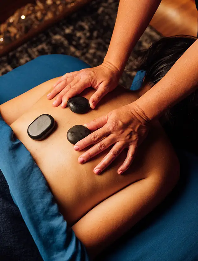An overhead view of a hot stone massage being performed on a client's back. The client is lying face down on a blue-covered table with three black stones placed on their back, and the masseuse's hands are applying pressure.