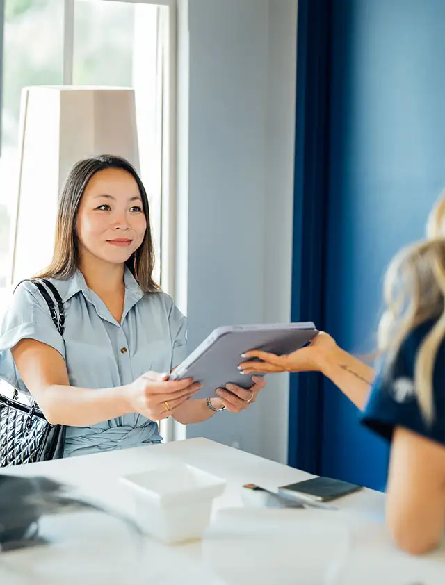 A smiling female client in a light blue shirt is handing documents across a white counter to an unseen person wearing dark blue scrubs.