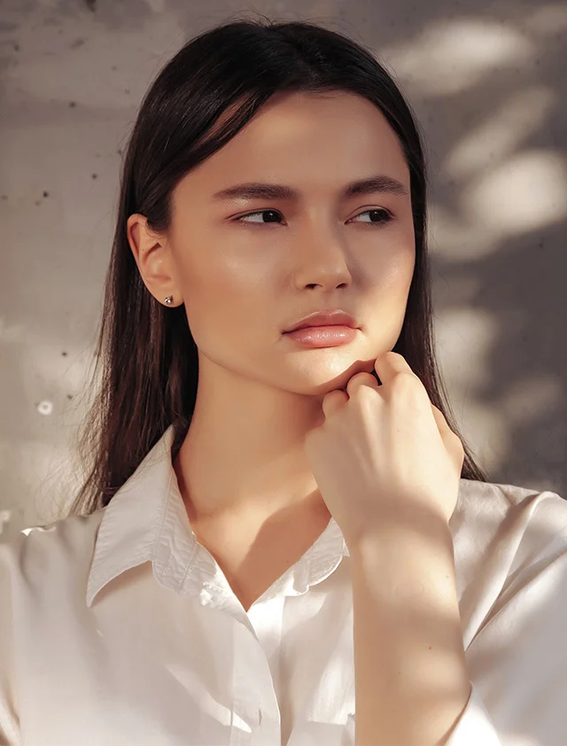 A woman with dark brown hair is seen from the mid-chest up against a textured light gray concrete wall. Her head is turned slightly to the right as she gazes into the distance. Dramatic lighting casts strong highlights on her face and hair, emphasizing her smooth, light-toned skin and subtle, natural makeup with rosy-pink lips. She appears bare-shouldered. - Microcurrent in Clarence, NY