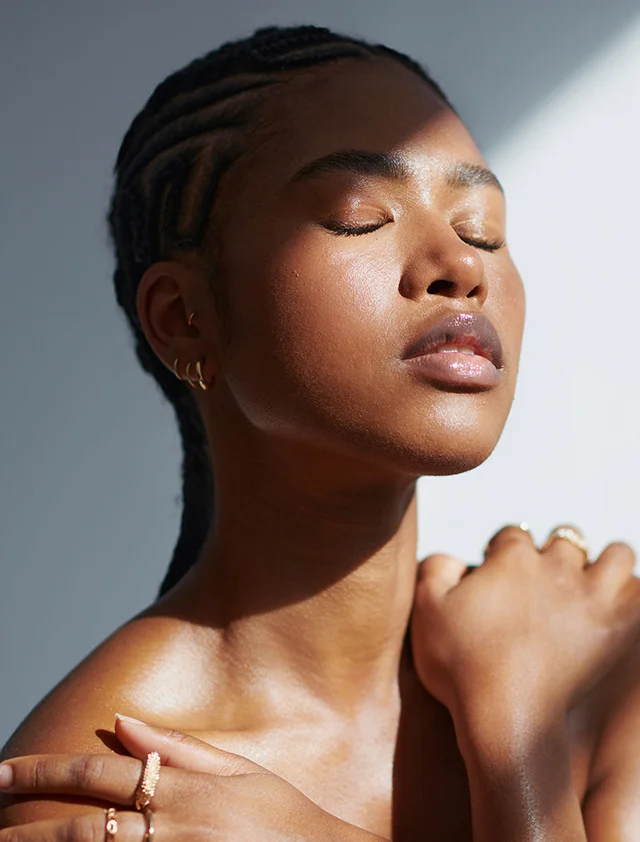 A young woman with glowing, dark skin and intricate cornrows stands in direct sunlight with her eyes closed and arms crossed over her bare shoulders. She is wearing several gold rings. - Microdermabrasion in Clarence, NY