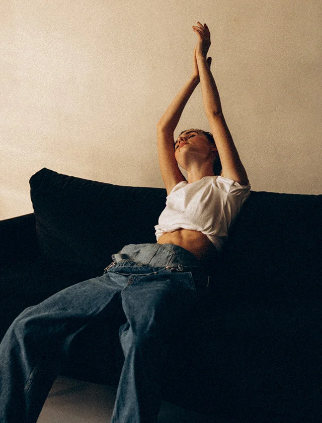 A woman sitting on a dark couch with her arms stretched up above her head. She is wearing a white crop top and wide-leg jeans with her eyes closed. - O Shot in Clarence, NY