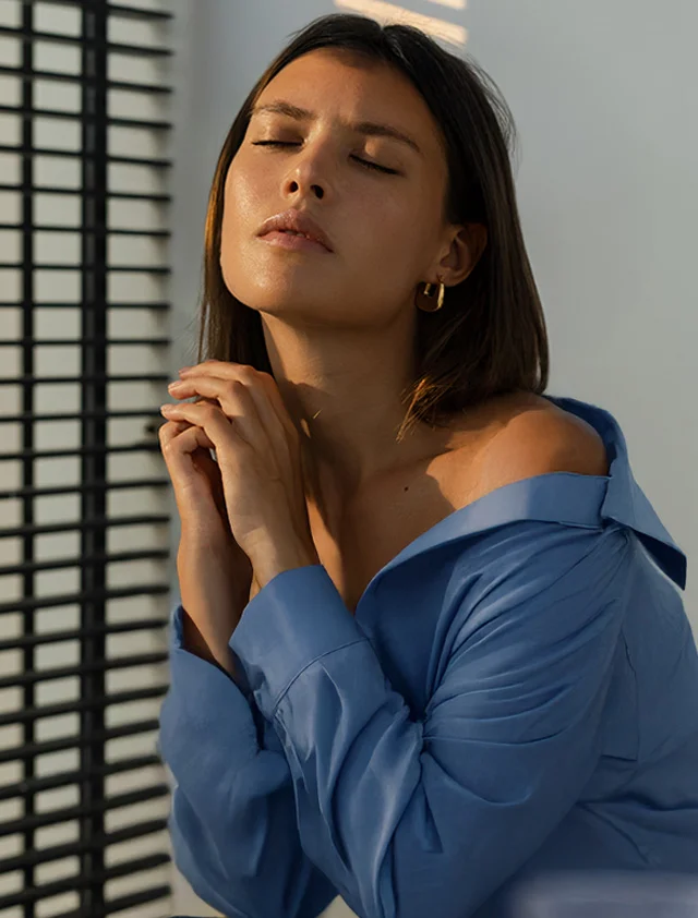 A woman with glowing skin, wearing a blue shirt, stands near window blinds with sun illuminating her face. Her eyes are closed and her hands are clasped near her chin. - Oxygen Facial in Clarence, NY