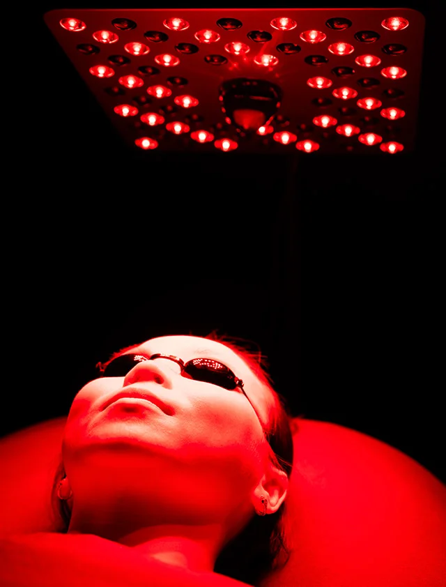 A person is lying down wearing small black protective eye coverings, illuminated by intense red light during a red light therapy session. - Red Light Therapy in Clarence, NY