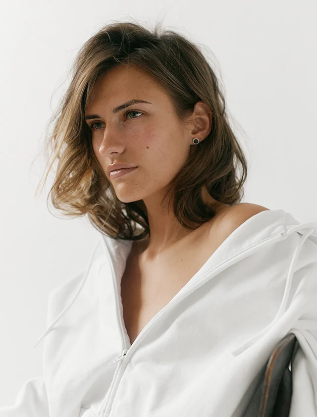 A young woman with brown, shoulder-length hair looks to the left, with her profile visible. She is wearing a white hoodie or top that is pulled down to expose her shoulder, and her hand is resting near her neck, gently touching her hair. She has a dark stud earring in her ear. The background is a plain, light color. - Sculptra in Clarence, NY