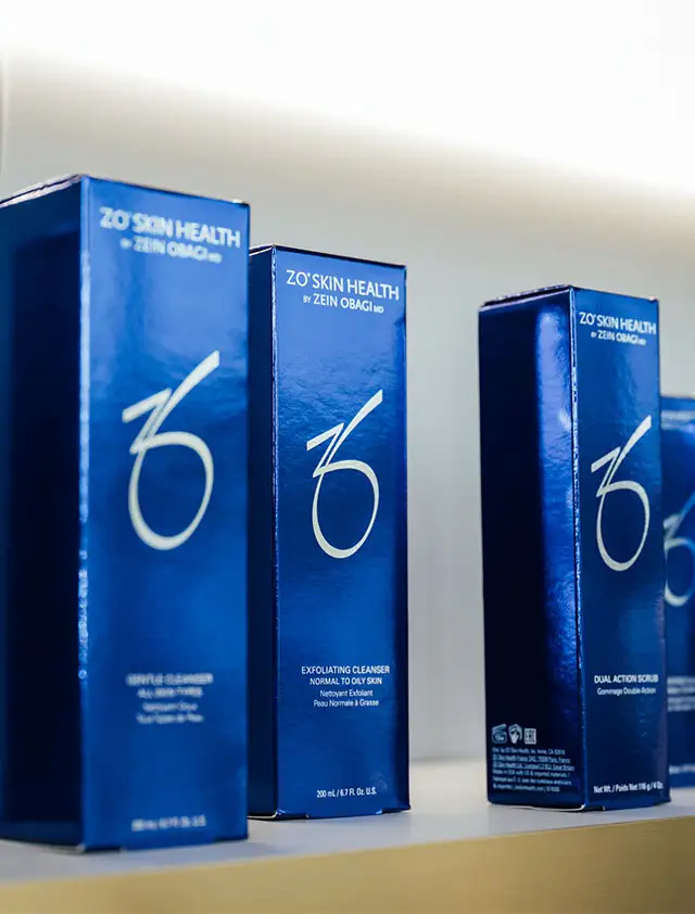 A row of tall, dark blue boxes of ZO Skin Health by Zein Obagi products are displayed on a white shelf. The boxes feature a white stylized "ZO" logo.
