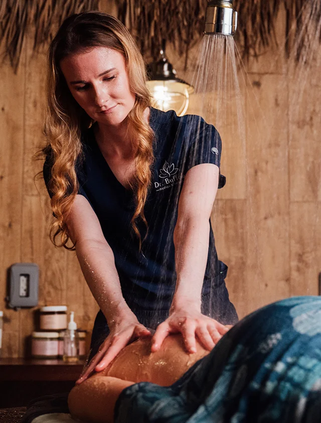 A blonde woman in a dark blue scrub top is giving a client a back massage in a dimly lit, wood-paneled spa room. - Vichy Vaginoplasty in Clarence, NY