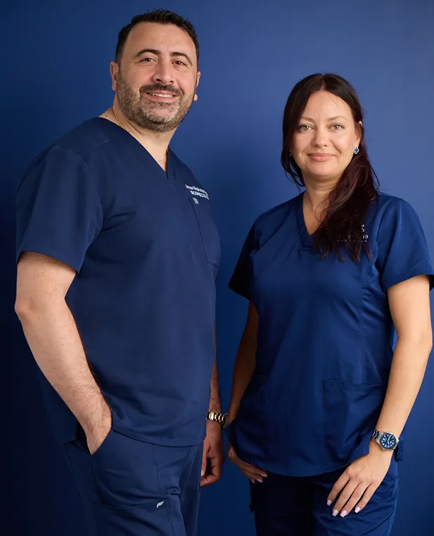 Dr. Kirakosyan, a man in a dark blue suit and tie, sits in a dark leather chair at a white desk, speaking to a blonde woman seated across from him. He is holding a pen and gesturing, with a white marble wall behind him.