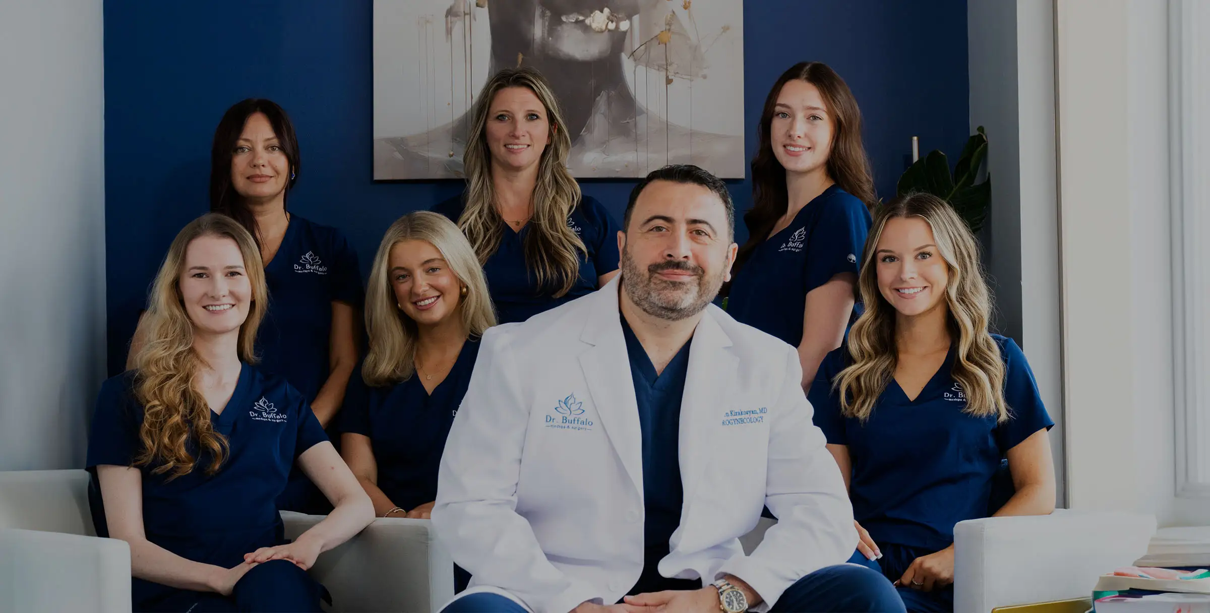 A group photo of Dr. Armen Kirakosyan and his team. Dr. Kirakosyan is seated in the center wearing a white lab coat over blue scrubs. Six women wearing matching dark blue scrubs stand or sit around him, all smiling. The background is a dark blue wall.